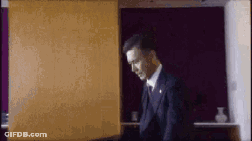 A Man In A Suit And Tie Is Standing In Front Of A Purple Wall In A Room . GIF