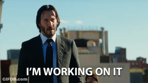 A Man In A Suit And Tie Is Standing In Front Of A Sign That Says I 'M Working On It GIF