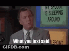 A Man In A Suit And Tie Is Standing In Front Of A Sign That Says My Spouse Is Sleeping Around . GIF