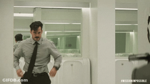 A Man In A Suit And Tie Is Standing In Front Of A Urinal In A Mission Impossible Scene GIF