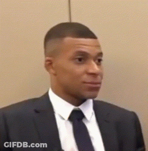 A Man In A Suit And Tie Is Standing In Front Of A Wall And Looking At The Camera . GIF