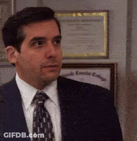 A Man In A Suit And Tie Is Standing In Front Of A Wall With Framed Pictures On It . GIF