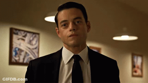 A Man In A Suit And Tie Is Standing In Front Of A Wall With Pictures On It GIF