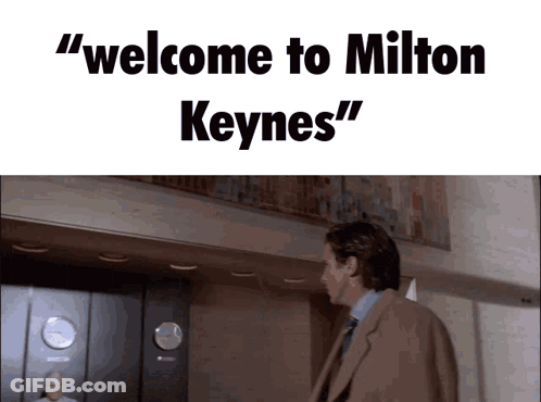 A Man In A Suit And Tie Is Standing In Front Of An Elevator With The Words " Welcome To Milton Keynes " Above Him GIF