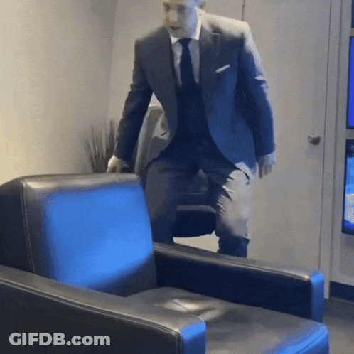 A Man In A Suit And Tie Is Standing Next To A Blue Chair GIF
