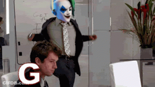 A Man In A Suit And Tie Is Standing Next To A Clown In A Mask With The Letter G Above Him GIF