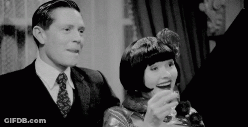 A Man In A Suit And Tie Is Standing Next To A Woman Holding A Glass Of Wine . GIF