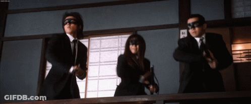 A Man In A Suit And Tie Is Standing Next To A Woman In A Mask And Holding A Gun GIF