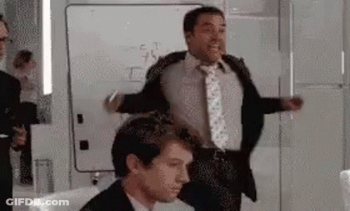A Man In A Suit And Tie Is Standing Next To Another Man In A Chair . GIF