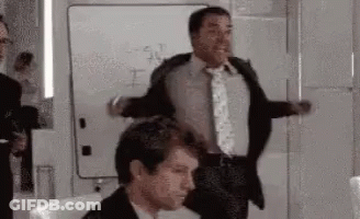 A Man In A Suit And Tie Is Standing Next To Another Man In A Chair . GIF