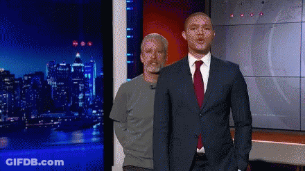 A Man In A Suit And Tie Is Standing Next To Another Man In A Grey Shirt GIF