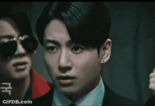A Man In A Suit And Tie Is Standing Next To Another Man Wearing Sunglasses And Earrings . GIF