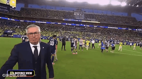 A Man In A Suit And Tie Is Standing On A Soccer Field With A Referee View Button On The Bottom GIF