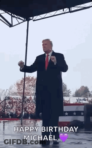 A Man In A Suit And Tie Is Standing On A Stage In Front Of A Crowd And Giving A Speech . GIF