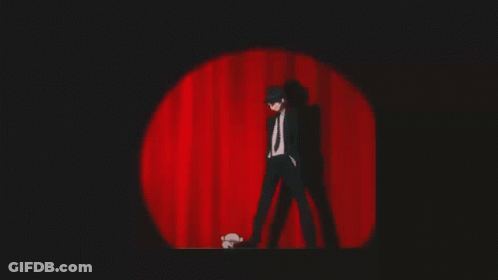 A Man In A Suit And Tie Is Standing On A Stage In Front Of A Red Curtain GIF