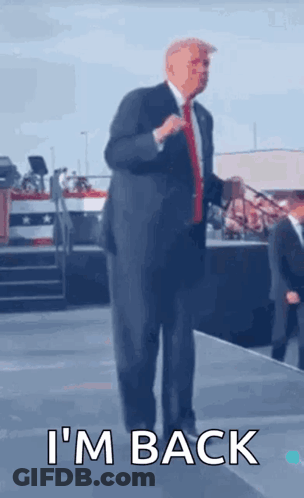 A Man In A Suit And Tie Is Standing On A Stage With The Words I 'M Back Written Below Him . GIF