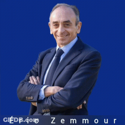 A Man In A Suit And Tie Is Standing With His Arms Crossed In Front Of A White Background With The Name Eric Zemmour On I GIF