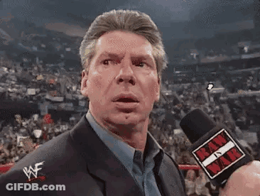 A Man In A Suit And Tie Is Talking Into A Microphone In A Stadium . GIF