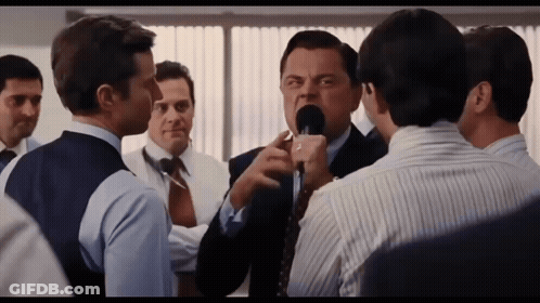 A Man In A Suit And Tie Is Talking Into A Microphone While Another Man Looks On GIF