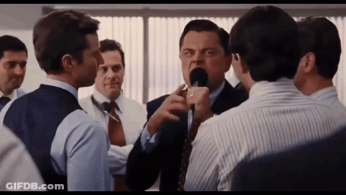 A Man In A Suit And Tie Is Talking Into A Microphone While Another Man Looks On GIF