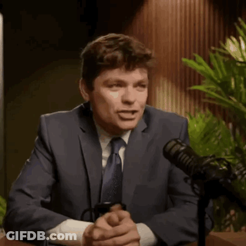 A Man In A Suit And Tie Is Talking Into A Microphone While Holding A Coffee Mug With The Letter Af On It . GIF