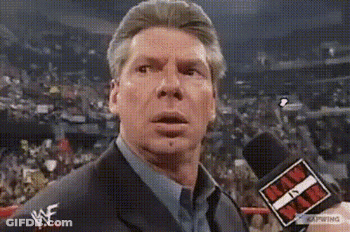 A Man In A Suit And Tie Is Talking Into A Microphone While Wearing A Wwe Logo On His Shirt . GIF