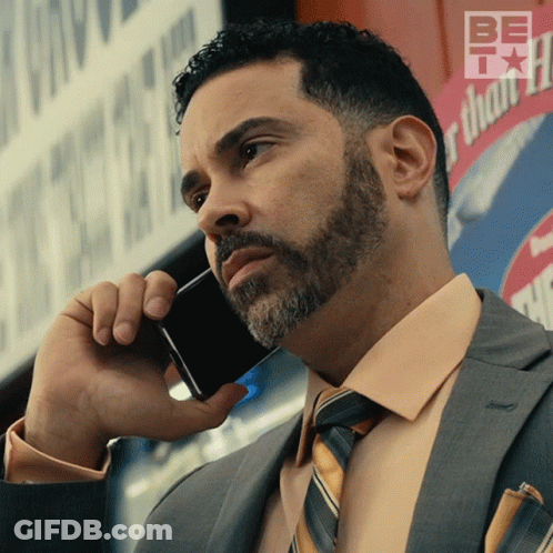 A Man In A Suit And Tie Is Talking On A Cell Phone In Front Of A Sign That Says Be Thankful GIF