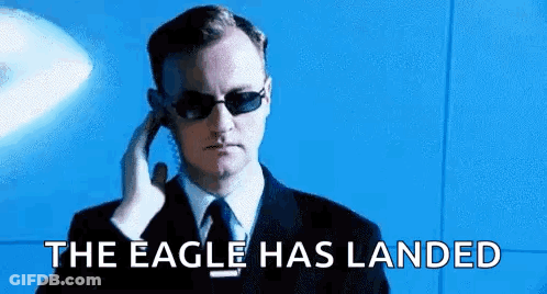 A Man In A Suit And Tie Is Talking On A Cell Phone With The Words `` The Eagle Has Landed '' Behind Him . GIF