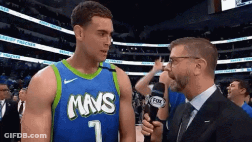 A Man In A Suit And Tie Is Talking To A Basketball Player In A Blue And Green Jersey . GIF