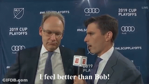 A Man In A Suit And Tie Is Talking To Another Man In Front Of A Banner That Says Mls Cup Playoffs GIF