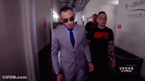 A Man In A Suit And Tie Is Walking Down A Hallway Next To A Man Wearing A Ufc Shirt . GIF
