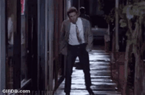 A Man In A Suit And Tie Is Walking Down A Narrow Alleyway At Night . GIF