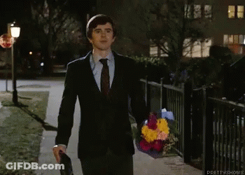 A Man In A Suit And Tie Is Walking Down A Sidewalk Holding A Bouquet Of Flowers .. GIF
