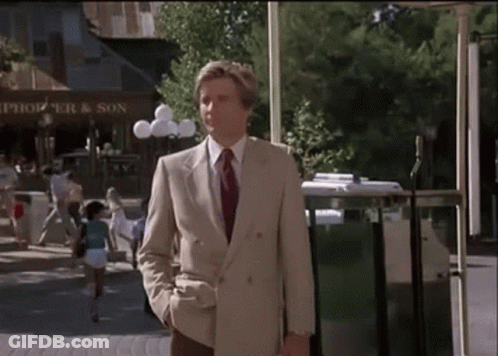 A Man In A Suit And Tie Is Walking Down A Street In Front Of A Store Called Thompson And Son GIF