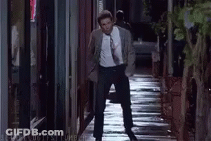 A Man In A Suit And Tie Is Walking Down A Wet Sidewalk At Night . GIF
