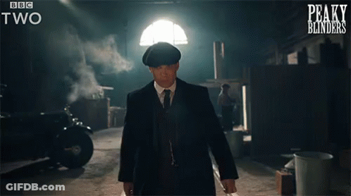 A Man In A Suit And Tie Is Walking In A Dark Room With Peaky Blinders Written On The Bottom Of The Screen GIF