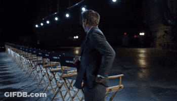 A Man In A Suit And Tie Is Walking In Front Of A Row Of Chairs GIF