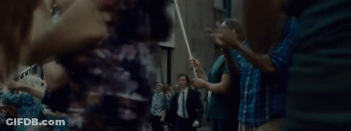A Man In A Suit And Tie Is Walking Through A Crowd Of People Holding A Stick . GIF