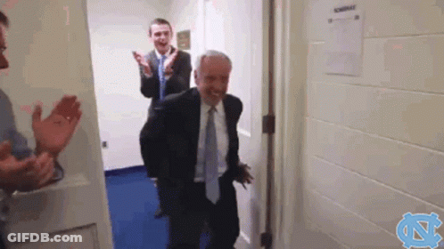 A Man In A Suit And Tie Is Walking Through A Doorway While Another Man Applauds . GIF