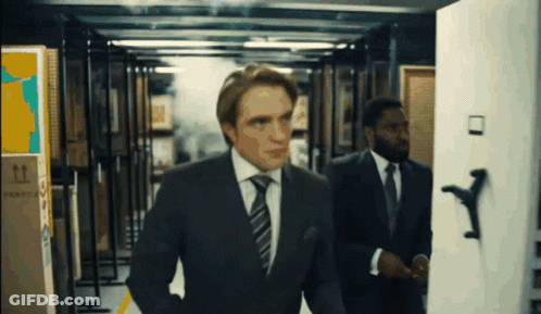 A Man In A Suit And Tie Is Walking Through A Hallway With A Box That Says Fragile On It GIF