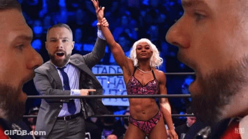 A Man In A Suit And Tie Points At A Woman In A Bikini In A Wrestling Ring GIF