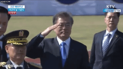 A Man In A Suit And Tie Salutes In Front Of A Live Ktv Screen GIF