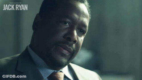 A Man In A Suit And Tie Says Okay In Front Of A Poster For Tom Clancy 'S Jack Ryan GIF