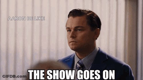 A Man In A Suit And Tie Says " The Show Goes On " In Front Of A White Curtain GIF
