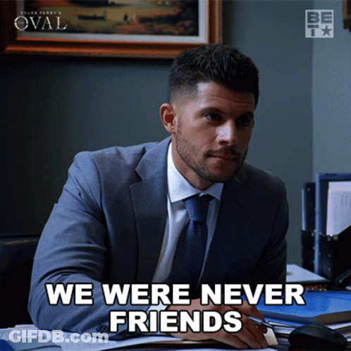 A Man In A Suit And Tie Sits At A Desk With The Words We Were Never Friends Below Him GIF