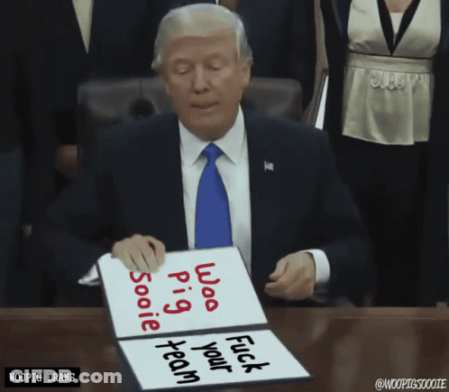 A Man In A Suit And Tie Sits At A Table Holding A Sign That Says " Whoo Pig " GIF