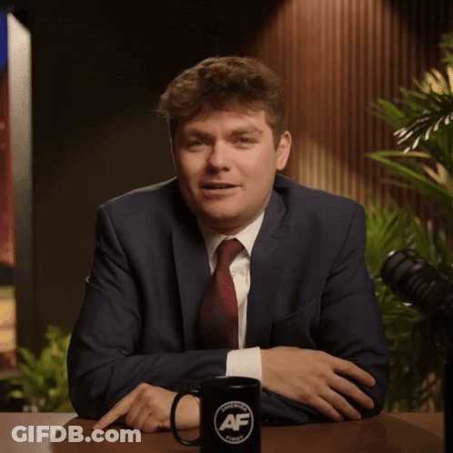 A Man In A Suit And Tie Sits At A Table Next To A Mug That Says Af On It GIF