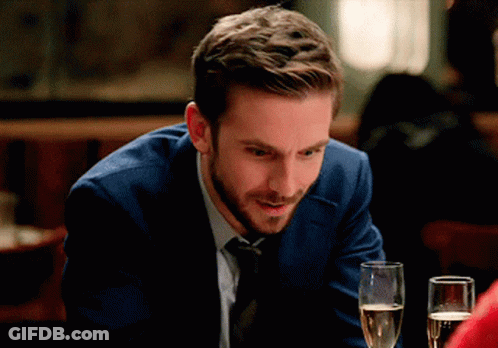 A Man In A Suit And Tie Sits At A Table With A Glass Of Champagne GIF