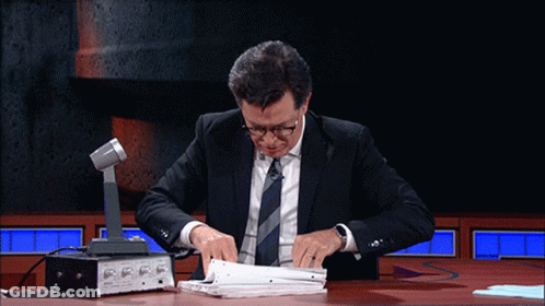 A Man In A Suit And Tie Sits At A Table With A Microphone GIF
