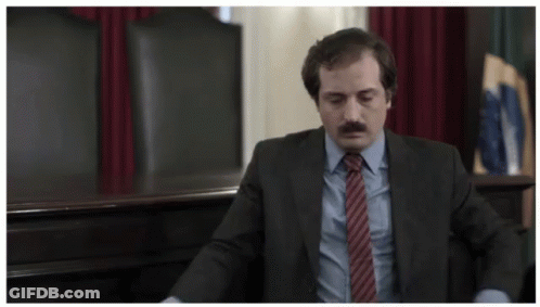 A Man In A Suit And Tie Sits In A Chair GIF